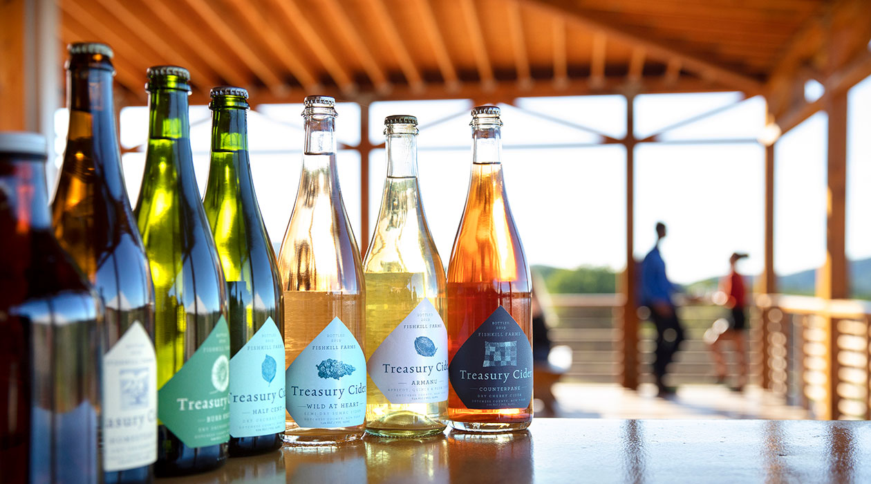 Six bottles of cider sit on a table on a deck with a couple out of focus in background