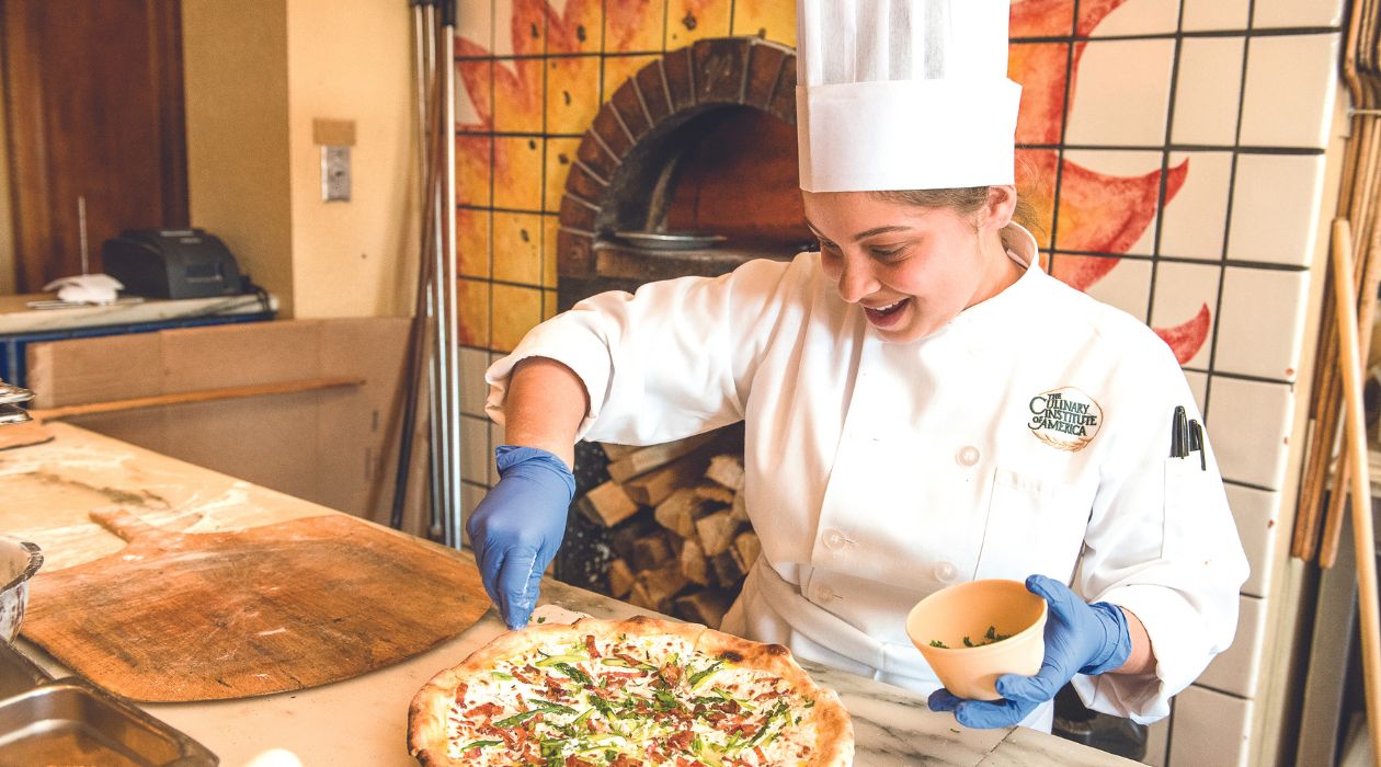 Culinary Institute of America Student in white uniform makes a pizza in front of a wood oven