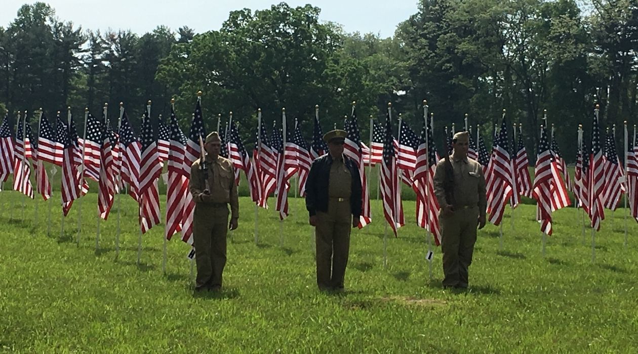 Three uniform clad soldiers stand in front of a field of American flags