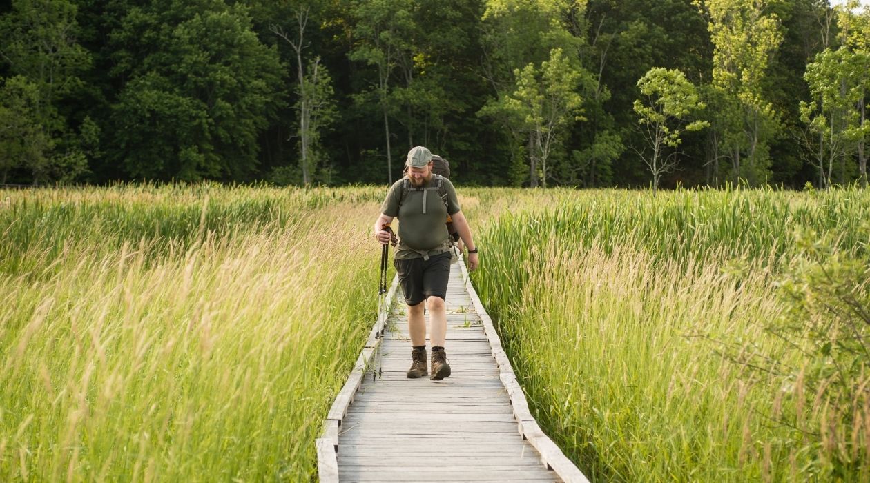 Man with backpack walking on boardwalk through overgrown swamp grass