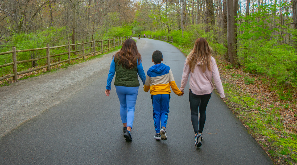 Three people walking are seen from behind on the Dutchess Rail Trail, a paved path weaving past green trees and grass. The person in the middle is a young boy, flanked by his mother and grandmother, holding hands