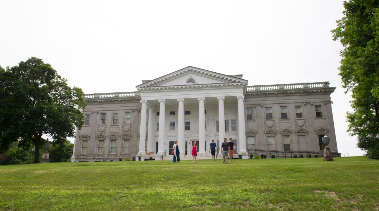 Staatsburgh State Historic Site, Staatsburg. A family walks along the grassy embankment in front of the 79 room stone mansion with its white columns and facade.