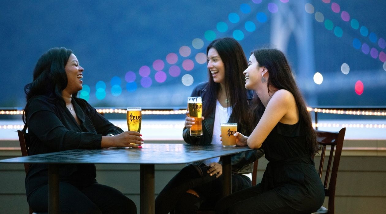 Three women sit around a table with bright beers on a rooftop with MidHudson bridge behind