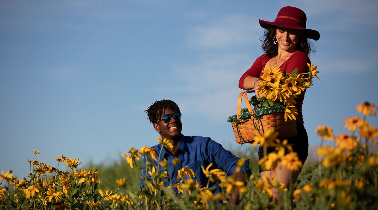 Man crouches in flower field while woman stands with bouquet
