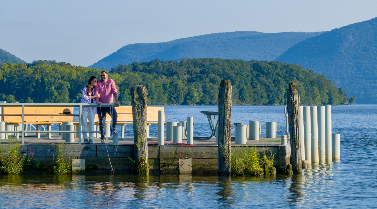 A couple is seen standing on a dock that juts out into blue waters of the Hudson River, with Hudson Highlands mountains seen behind them in the distance